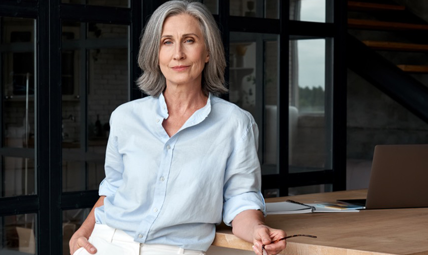 Older woman leaning against a desk with classes in her hand.