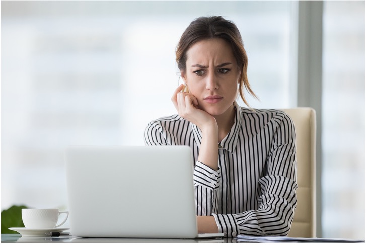 A woman sitting at a desk looking at a laptop with a concerned expression.